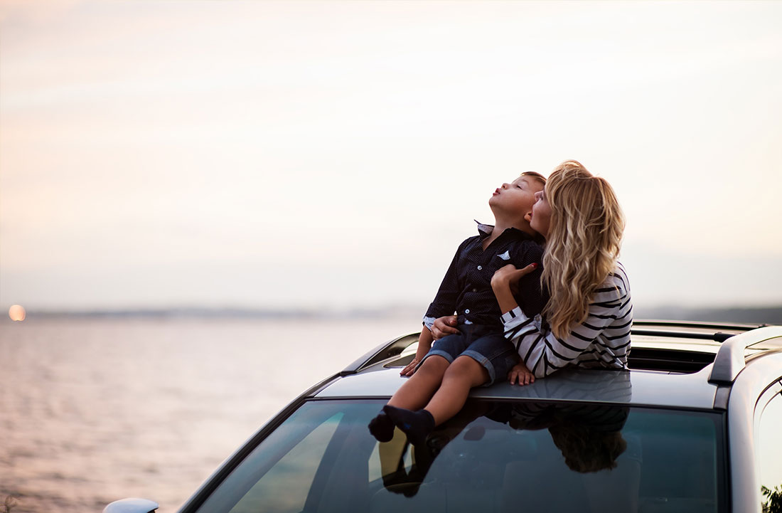 boy sitting on top of a car and mom holding him and looking up